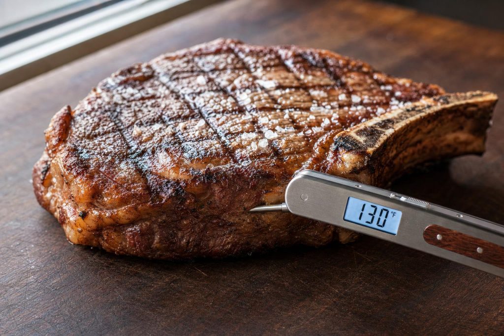 A slim brushed-stainless folding instant-read thermometer inserted into a thick bone-in ribeye steak on a dark walnut cutting board with the display reading 130°F.