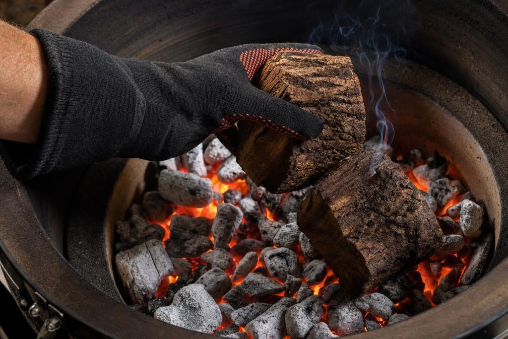 A gloved hand placing two irregular hickory wood chunks onto a tight pile of glowing red-orange charcoal coals inside a ceramic kamado grill.